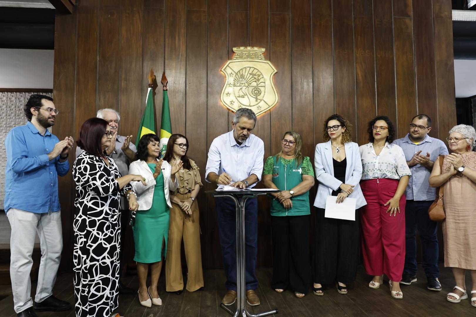 Elmano de Freitas, em camisa azul clara, assina um documento em um pódio durante cerimônia, cercado por um grupo diverso de pessoas em um salão com painéis de madeira e brasão oficial.