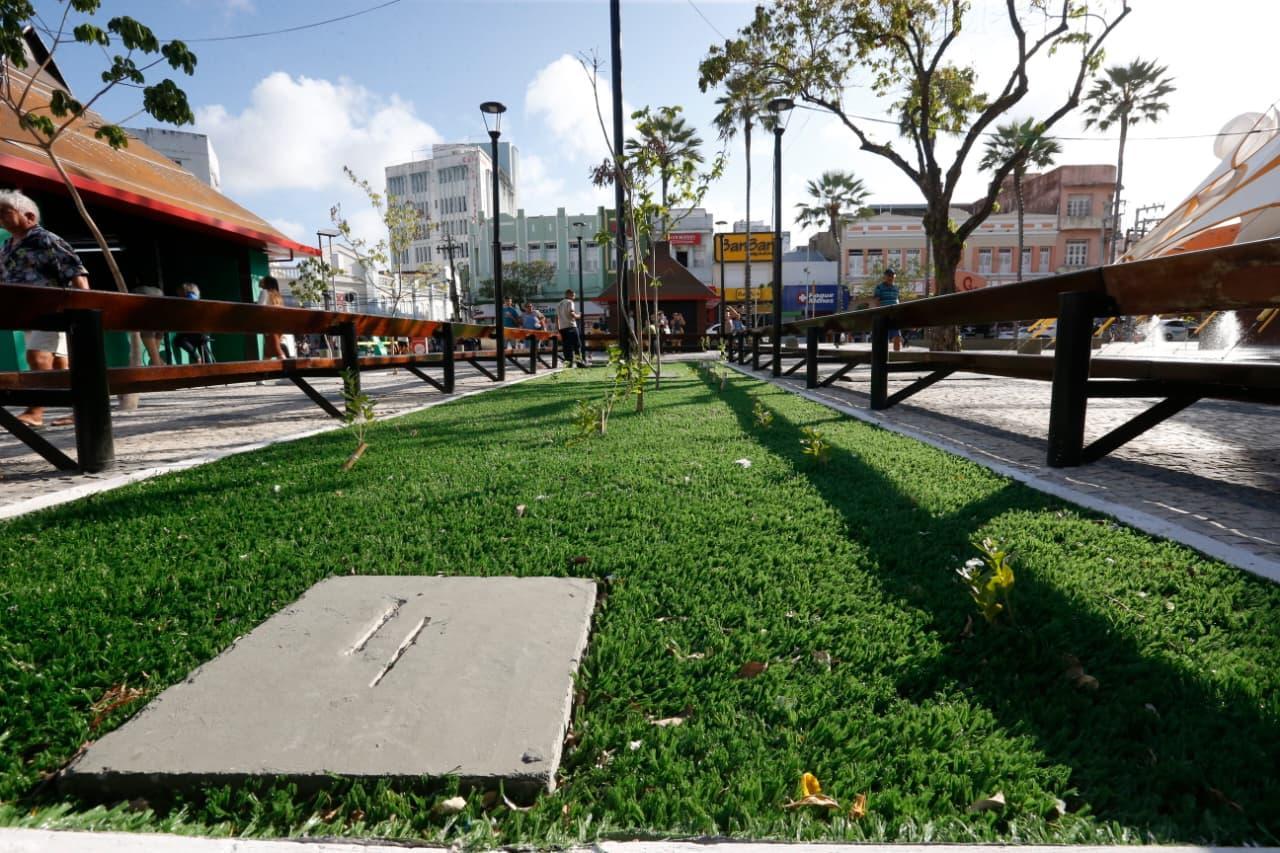 Canteiro de grama sintética na Praça do Ferreira, em Fortaleza, com uma placa de concreto no primeiro plano, emoldurado por longos bancos de madeira em uma praça movimentada. Edifícios históricos e vegetação urbana são visíveis ao longe, sob a luz do sol.