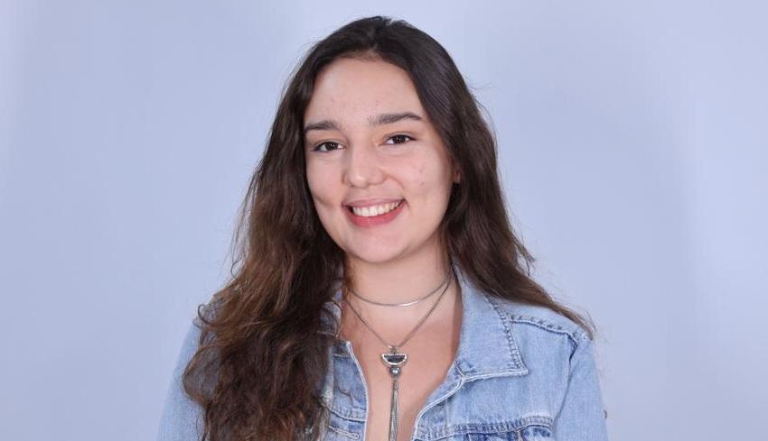 Mulher jovem sorrindo, de cabelos longos e castanhos, usando jaqueta jeans clara e colares, fotografada em fundo neutro claro, em retrato de estúdio.