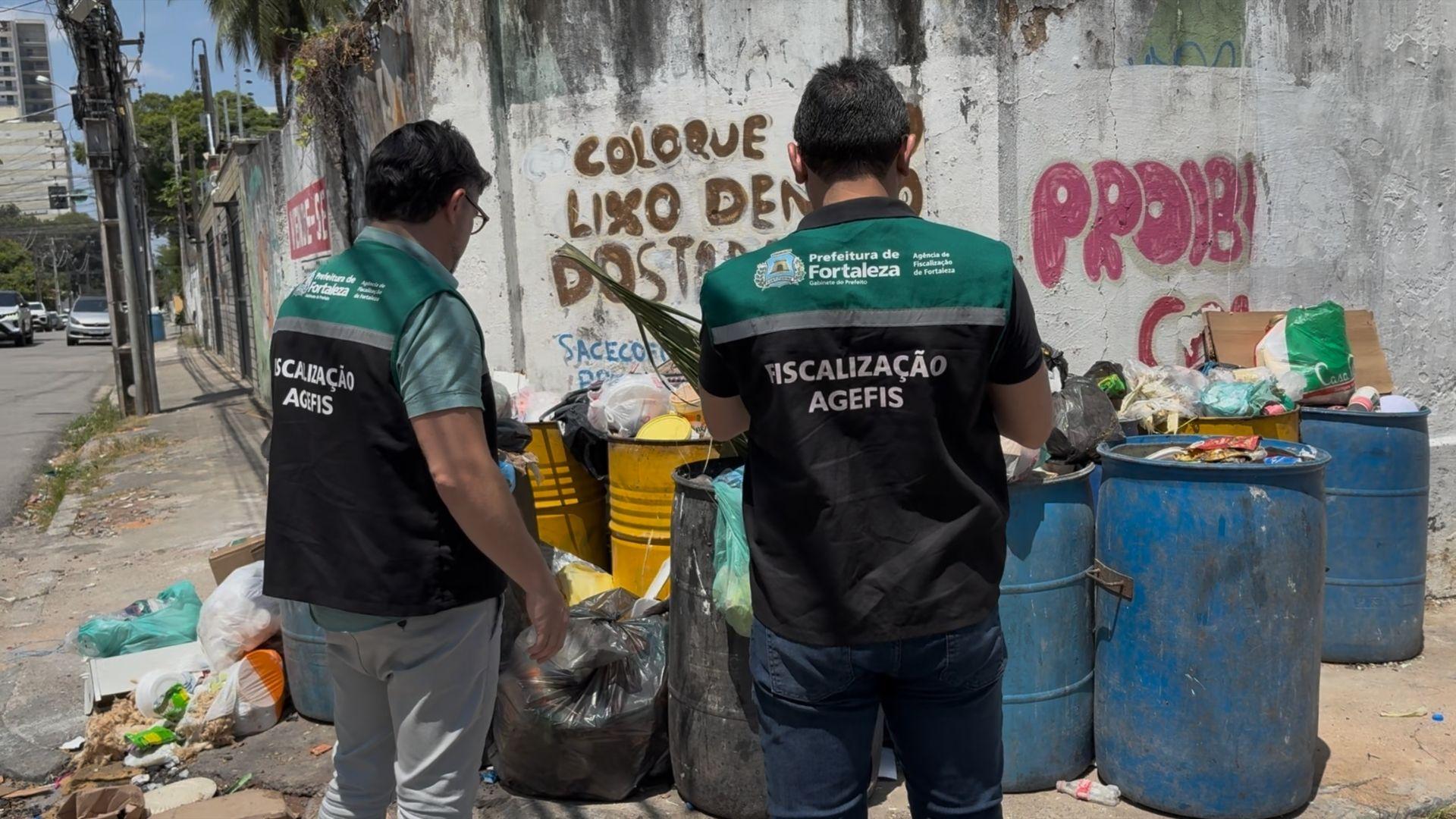 Dois fiscais da AGEFIS, Agência de Fiscalização de Fortaleza, inspecionam um ponto de descarte irregular de lixo em uma via urbana, com tambores cheios de resíduos e grafites na parede sobre descarte.
