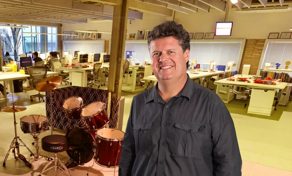 Homem em pé, sorrindo, em primeiro plano de um escritório aberto. Ao fundo, aparecem mesas de trabalho ocupadas por equipes e uma bateria montada como elemento decorativo no ambiente da agência.