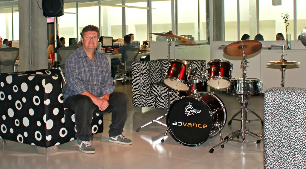 Homem sentado em um ambiente de escritório moderno, ao lado de uma bateria com o nome “Advance” no bumbo. Ao fundo, é possível ver colaboradores trabalhando em mesas, com computadores, em um espaço amplo e iluminado.