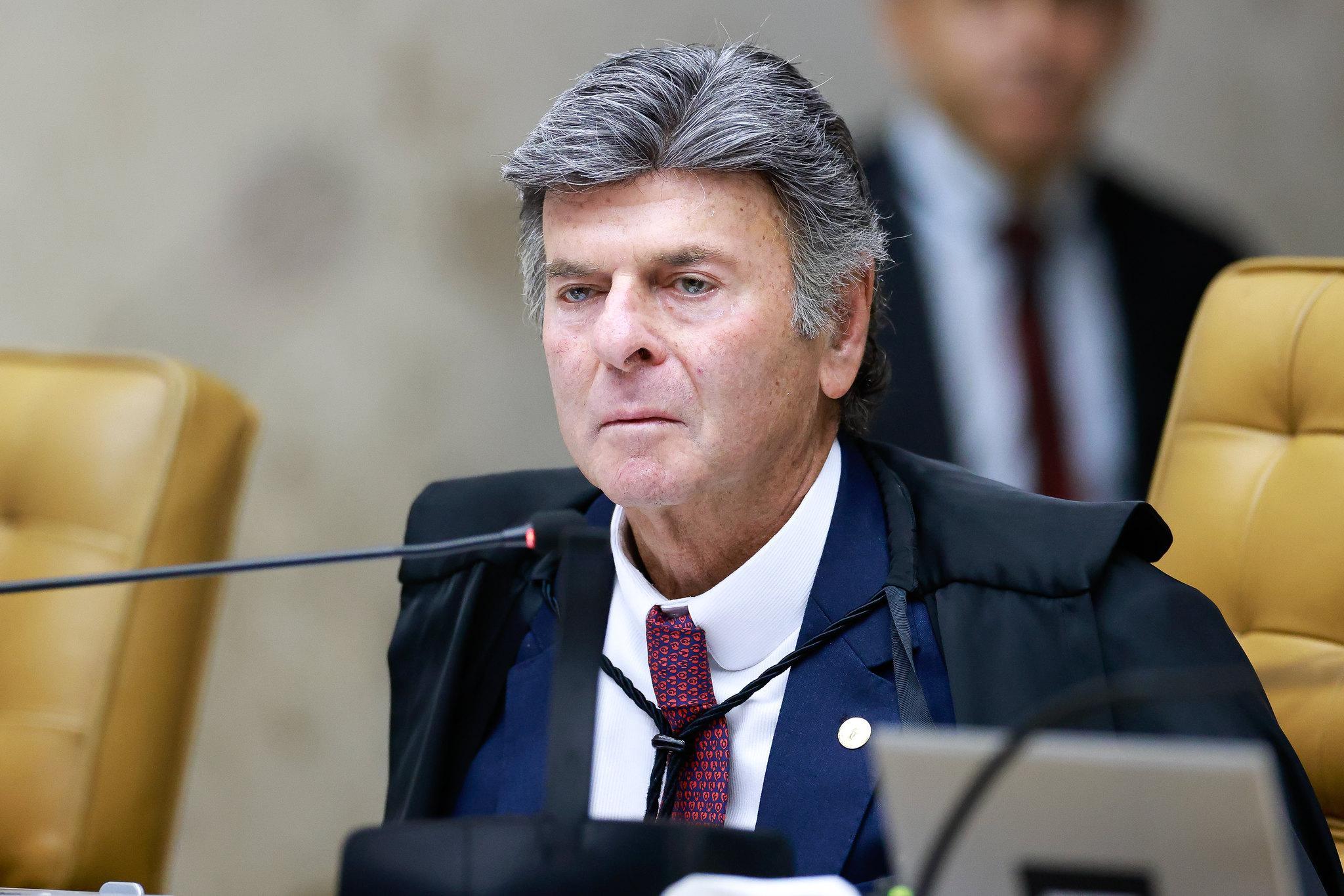 Fux usando toga preta está sentada em uma cadeira estofada em uma sala de tribunal. À frente dela há um microfone e um computador. Ao fundo, aparece outra pessoa desfocada, também em trajes formais. Imagem usada em matéria sobre ministro suspender bloqueio de Bets para beneficiários do Bolsa Família.