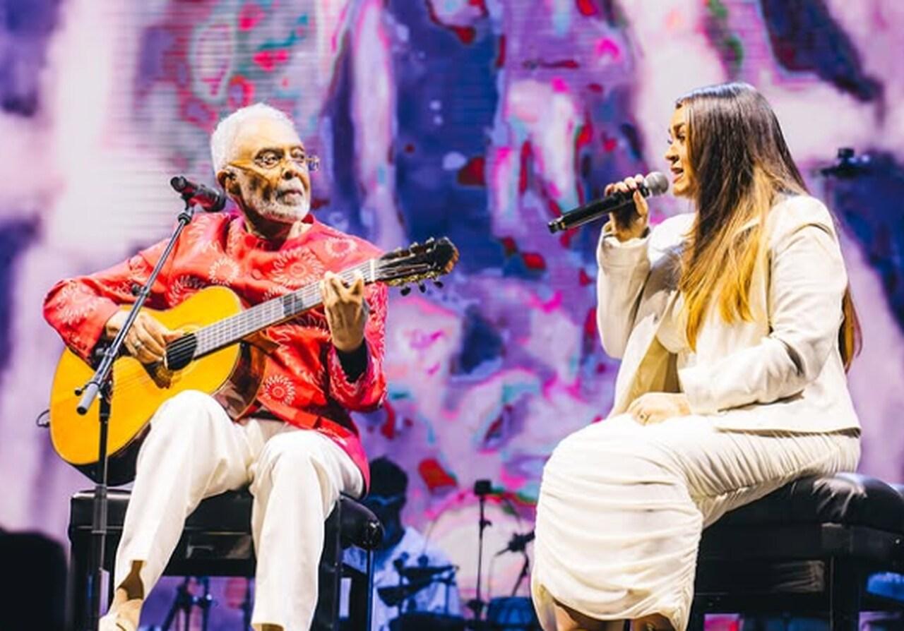 Gilberto e Preta sentados lado a lado em um palco, ele com um violão e ambos com um microfone. Ele, à esquerda, é um homem preto de cabelos brancos e óculos, com roupa rosa de manga longa e calça branca. Ela, à direita, é uma mulher preta com cabelos lisos longos castanhos, de pontas loiras, e roupa longa branca.