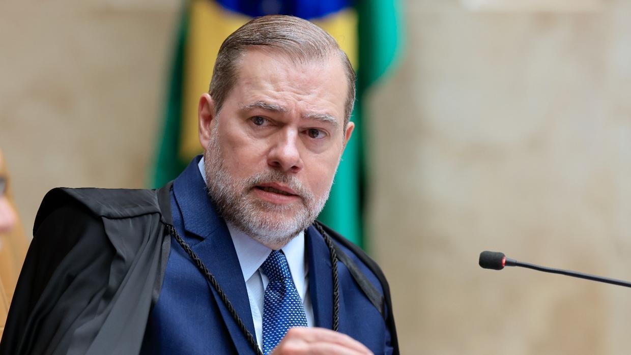 Na imagem, fotografia em plano médio do ministro Dias Toffoli, do Supremo Tribunal Federal (STF). Ele é um homem de pele clara, cabelos castanhos curtos e barba grisalha aparada. Veste uma toga preta sobre um terno azul-escuro, camisa social clara e gravata azul com pequenos detalhes. O ministro está levemente inclinado para a frente, com uma expressão séria e a boca entreaberta, como se estivesse falando. À sua frente, no canto inferior direito, aparece parte de um microfone preto. Ao fundo, de forma desfocada, vê-se a bandeira do Brasil.