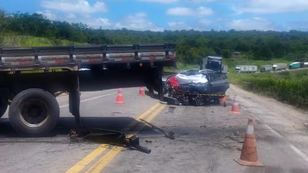 Uma imagem de um cenário de acidente automobilístico em uma rodovia de pista simples, sob luz do dia. Em primeiro plano, à esquerda, vê-se a lateral traseira de uma carreta de carga, com um pneu de grande porte e parte da carroceria de madeira. Ao centro, um carro de passeio azul escuro está completamente destruído após a colisão, com a parte frontal esmagada e o teto retorcido. Diversos cones de sinalização laranjas estão espalhados pela pista ao redor dos veículos para isolar a área, e uma fita amarela de isolamento atravessa a frente do carro. Ao fundo, há uma vegetação densa e verde, além de outros caminhões parados no acostamento da estrada sob um céu com poucas nuvens.