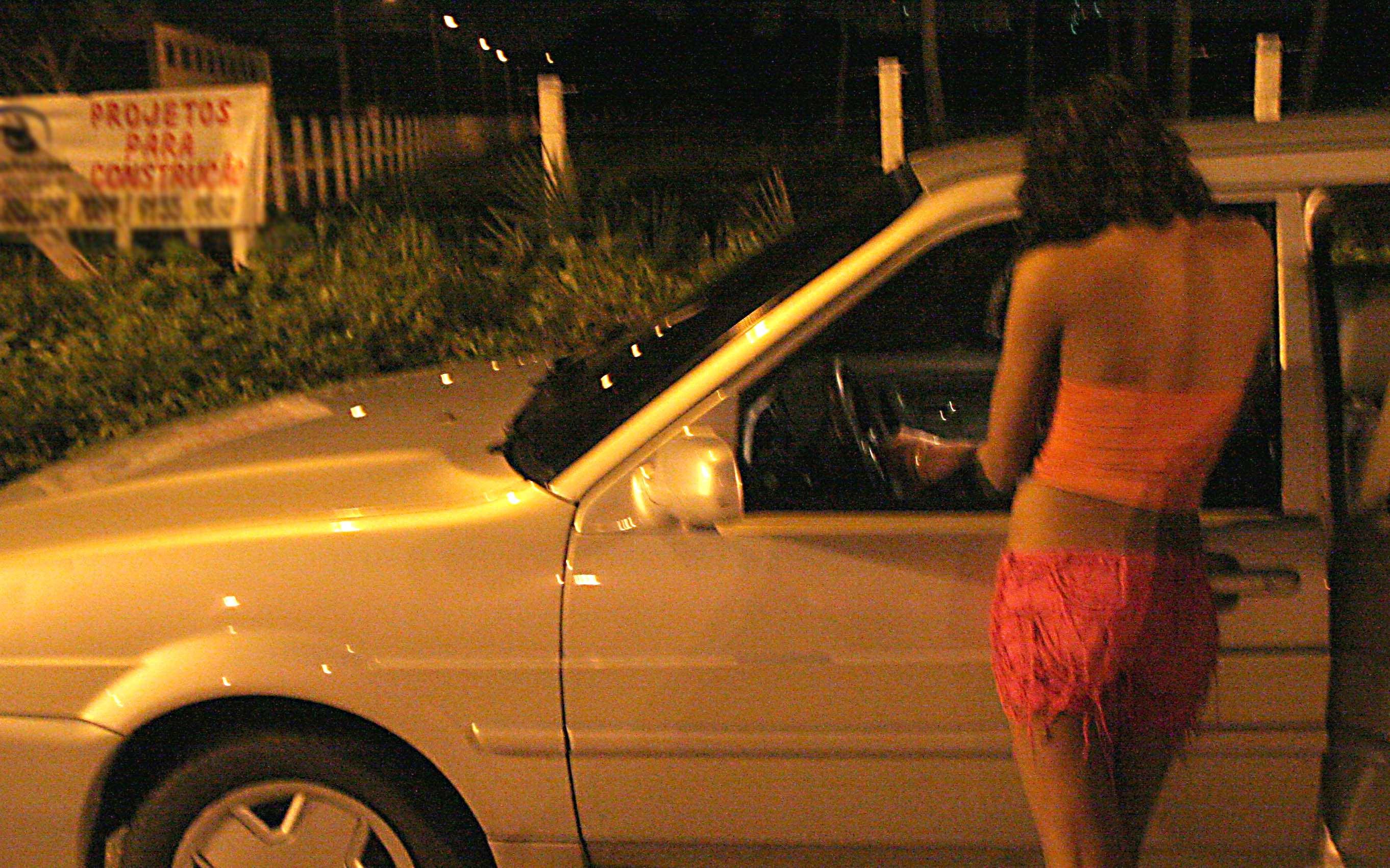 Fotografia noturna em plano médio de uma mulher de costas, em pé ao lado de um carro prata estacionado. Ela veste um top laranja e um short curto rosa com franjas. A mulher está com o braço estendido em direção ao interior do veículo através da janela aberta do motorista. O ambiente é pouco iluminado por uma luz amarelada. Ao fundo, há vegetação rasteira e uma placa branca com letras vermelhas onde se lê 