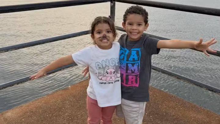 Duas crianças estão de pé lado a lado em um deck de concreto à beira da água, cercado por um guarda‑corpo metálico. As duas esticam os braços para os lados, como se imitassem asas. A criança à esquerda usa camiseta branca com um desenho de personagem, calça rosa e tênis claros. A criança à direita veste camiseta cinza com letras coloridas estampadas, bermuda clara e tênis pretos. Ao fundo, a água está tranquila, sugerindo um fim de tarde.