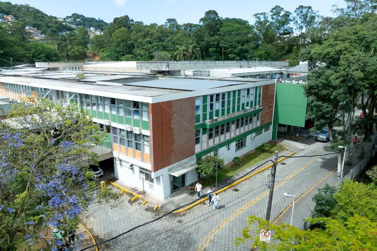 Vista aérea de um prédio grande e baixo, com vários blocos interligados, janelas amplas e detalhes nas cores verde, branco e tijolo aparente. A construção está cercada por muitas árvores e vegetação, com uma rua de paralelepípedos passando em frente. Algumas pessoas caminham pela calçada e há carros estacionados ao redor. O cenário parece ser de um campus ou complexo institucional localizado em uma área arborizada.