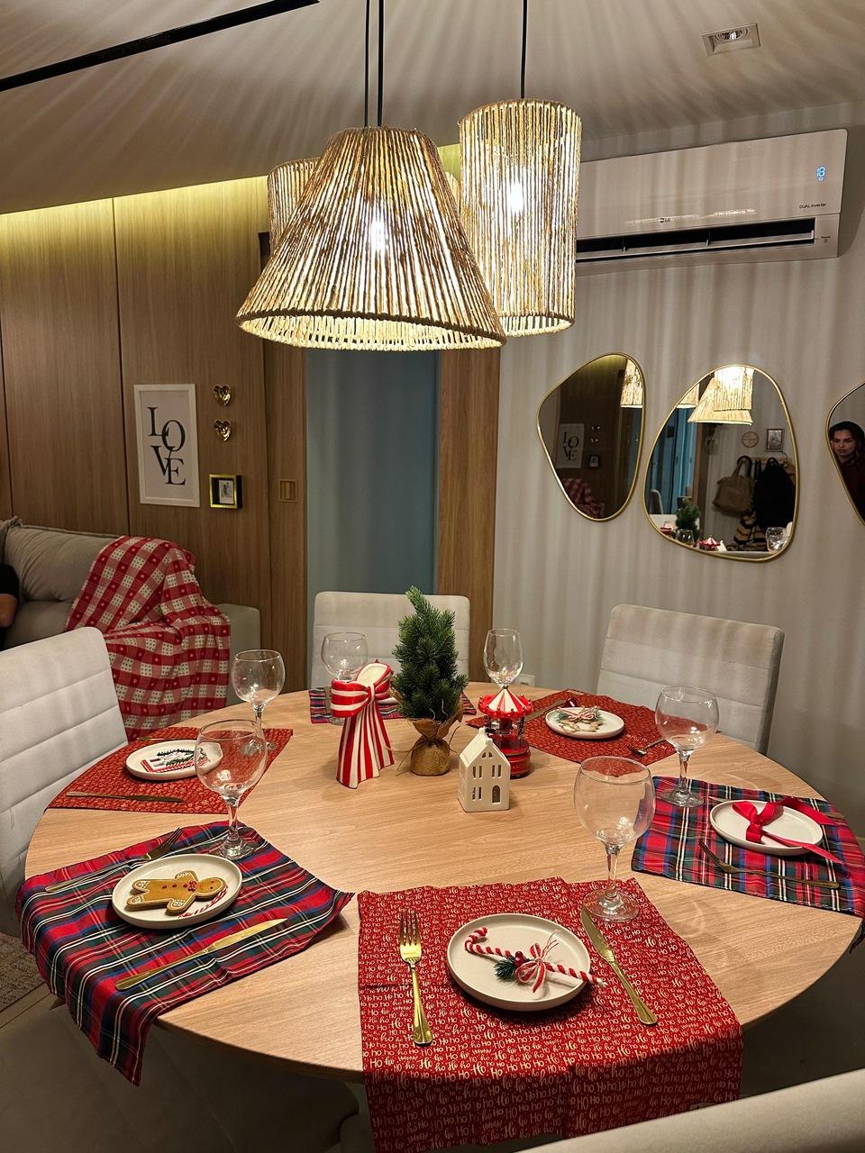 Uma fotografia colorida, capturada de um ângulo levemente elevado, mostra uma mesa de jantar redonda de madeira clara decorada com temática natalina. A cena transmite uma atmosfera aconchegante e acolhedora, com iluminação quente vinda de pendentes rústicos.