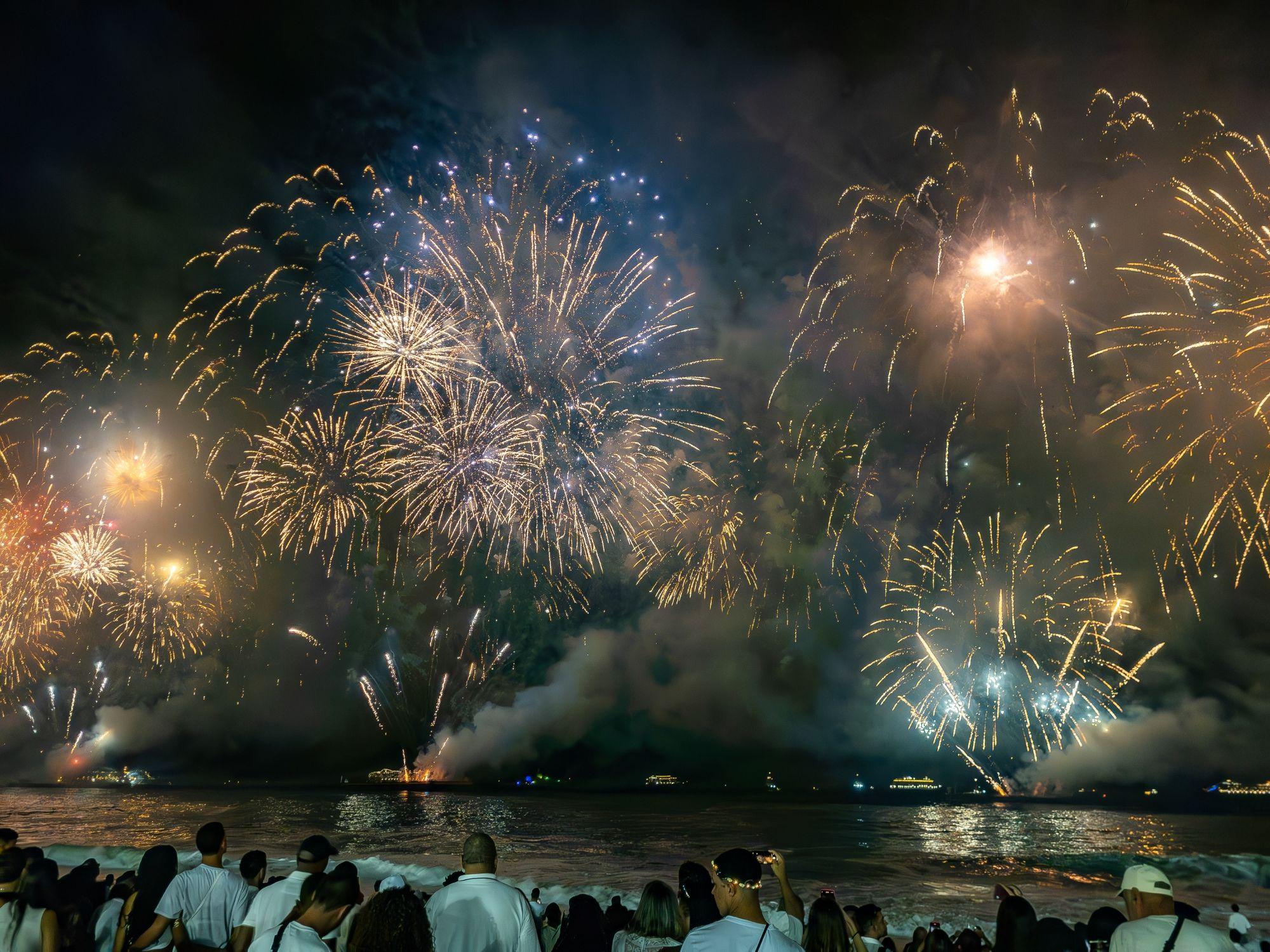Pessoas vestidas de branco observam um grandioso espetáculo de fogos de artifício em uma praia durante a noite de réveillon.