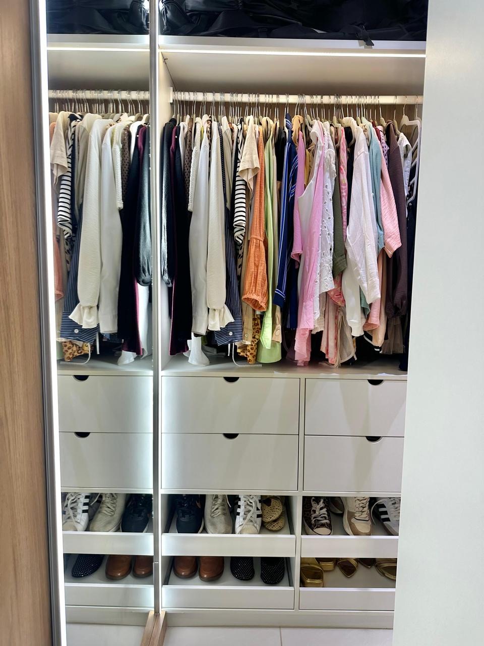 Interior de um guarda-roupa planejado com iluminação em LED, exibindo roupas penduradas e organizadas por cores acima de seis gavetas brancas. Na base, prateleiras deslizantes acomodam diversos pares de sapatos alinhados de forma a otimizar o espaço.