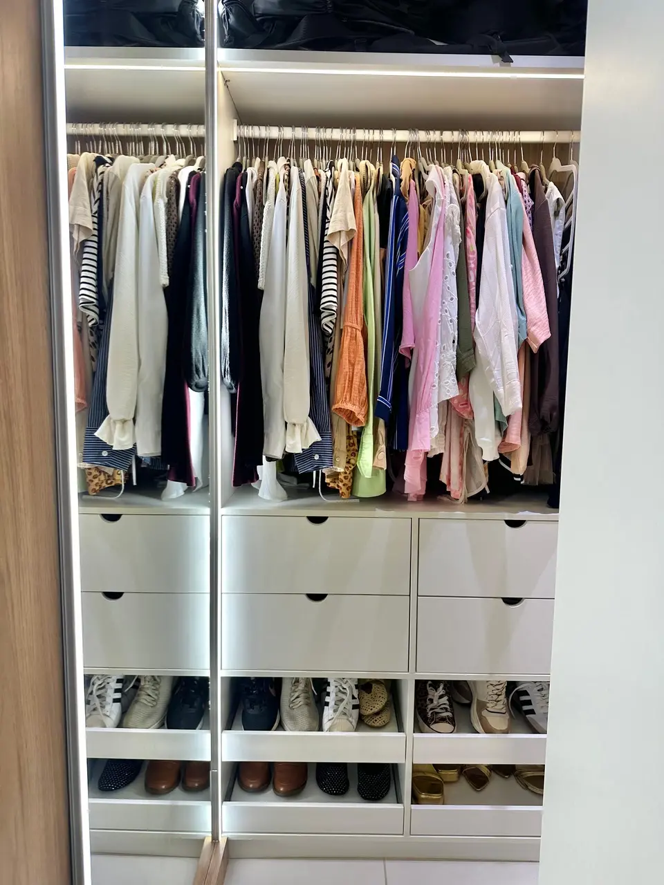 Interior de um guarda-roupa planejado com iluminação em LED, exibindo roupas penduradas e organizadas por cores acima de seis gavetas brancas. Na base, prateleiras deslizantes acomodam diversos pares de sapatos alinhados de forma a otimizar o espaço.