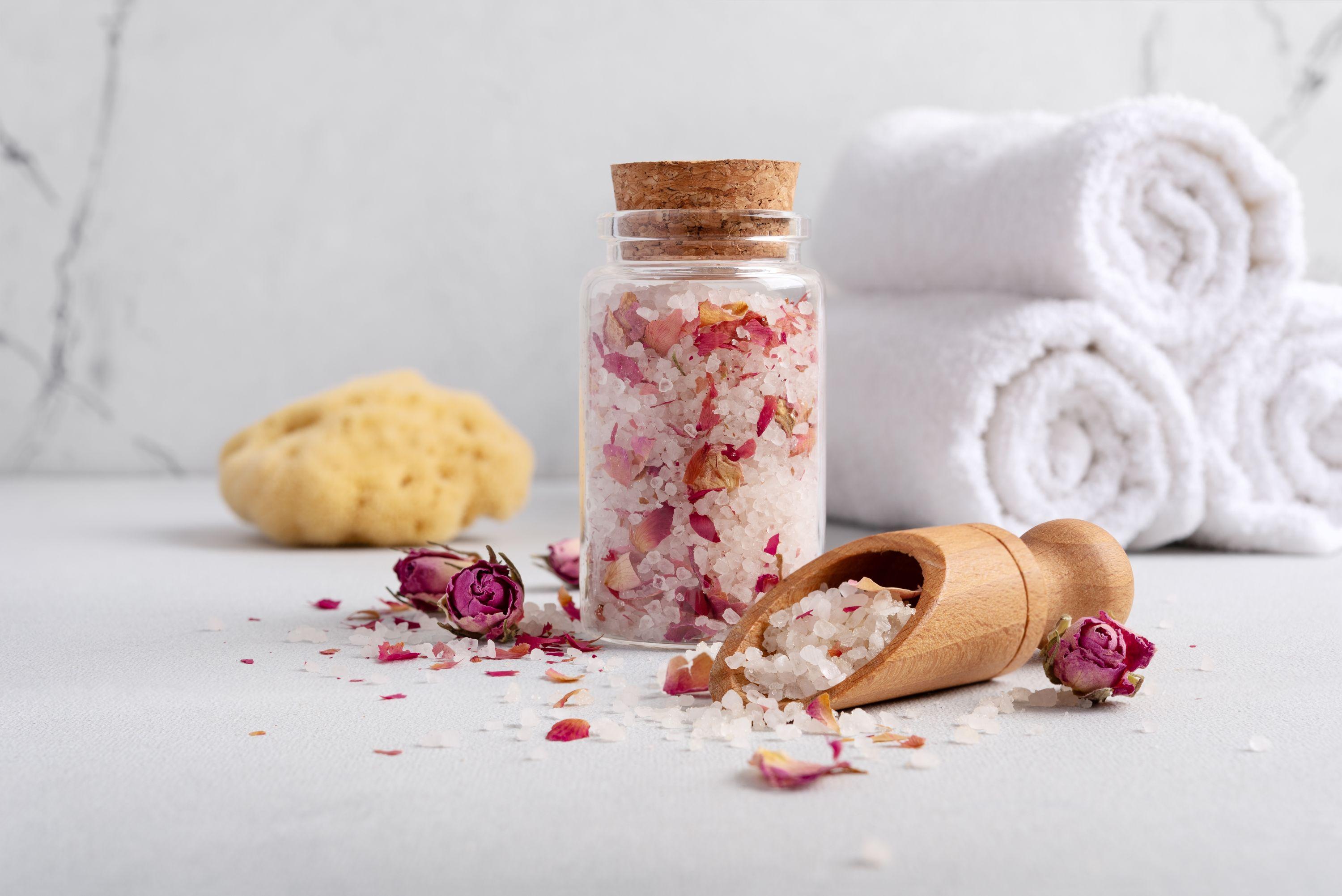Uma garrafa de vidro com sais de banho de rosa e brancos, uma colher de madeira, pétalas de rosa secas, toalhas brancas enroladas e uma esponja natural em uma superfície clara.