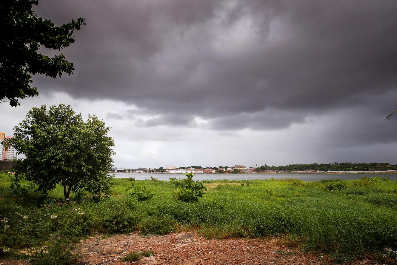 Paisagem de um lago ou rio com céu dramaticamente nublado, vegetação verde em primeiro plano e construções distantes na margem.