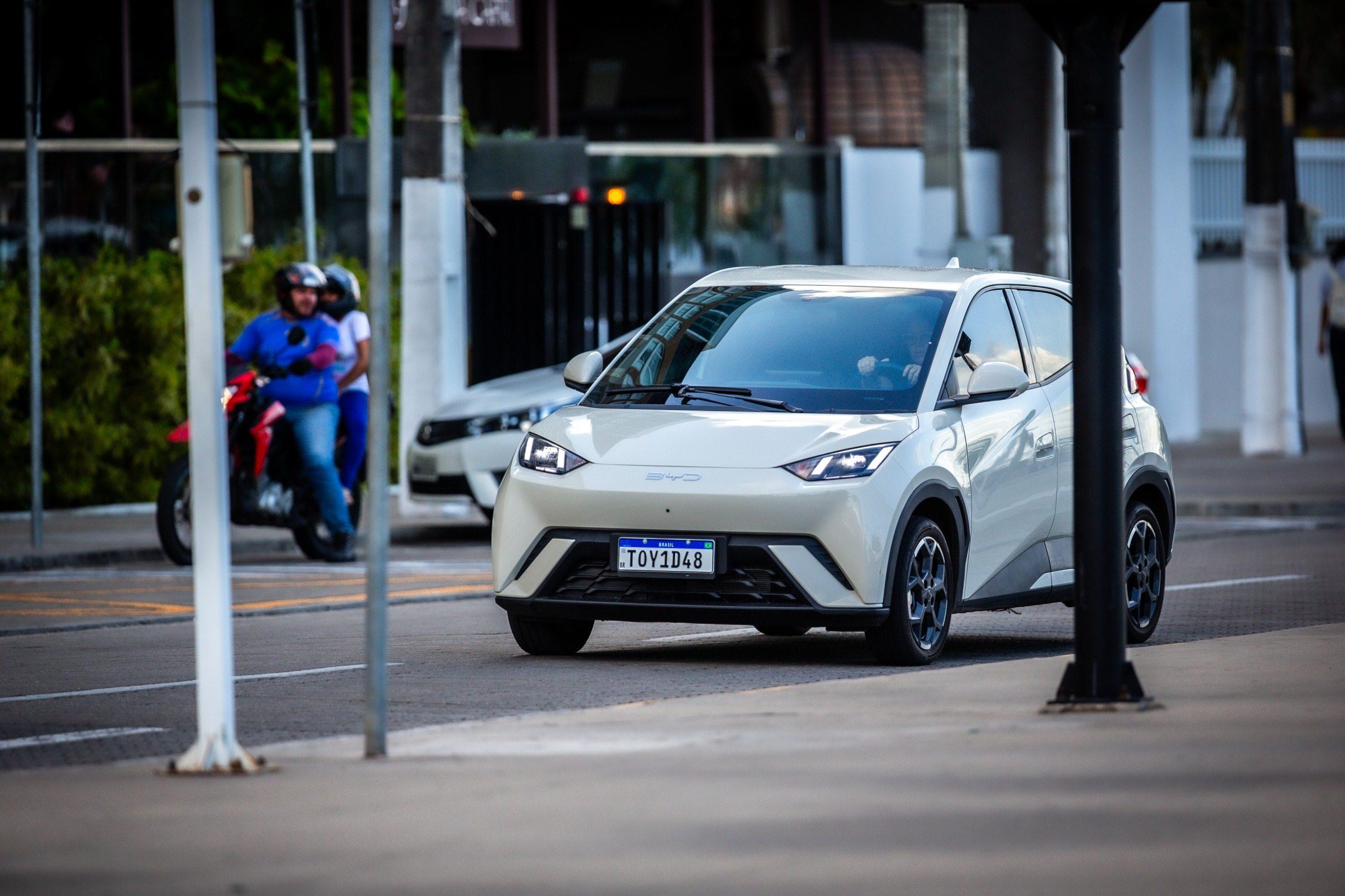 Fotografia em plano médio de um carro elétrico compacto BYD Dolphin Mini, na cor branco perolado, trafegando por uma rua urbana. O veículo está em foco no centro da imagem, apresentando design moderno com faróis afilados e a placa Mercosul TOY1D48. Ao fundo, à esquerda, um motociclista e uma passageira em uma moto vermelha aparecem levemente desfocados, assim como os elementos da calçada e postes em primeiro plano, criando profundidade e destaque para o automóvel principal.