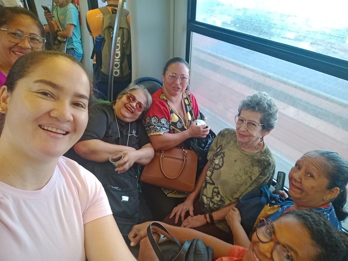 Na imagem, uma selfie em ângulo levemente inclinado captura um grupo alegre de sete mulheres de diversas idades dentro de um vagão de metrô. Elas estão sorridentes, algumas sentadas e outras em pé, transmitindo um clima de amizade e descontração. Ao fundo, através da janela do trem, vê-se o movimento do lado de fora e outros passageiros. A iluminação é natural, vinda das janelas do vagão.