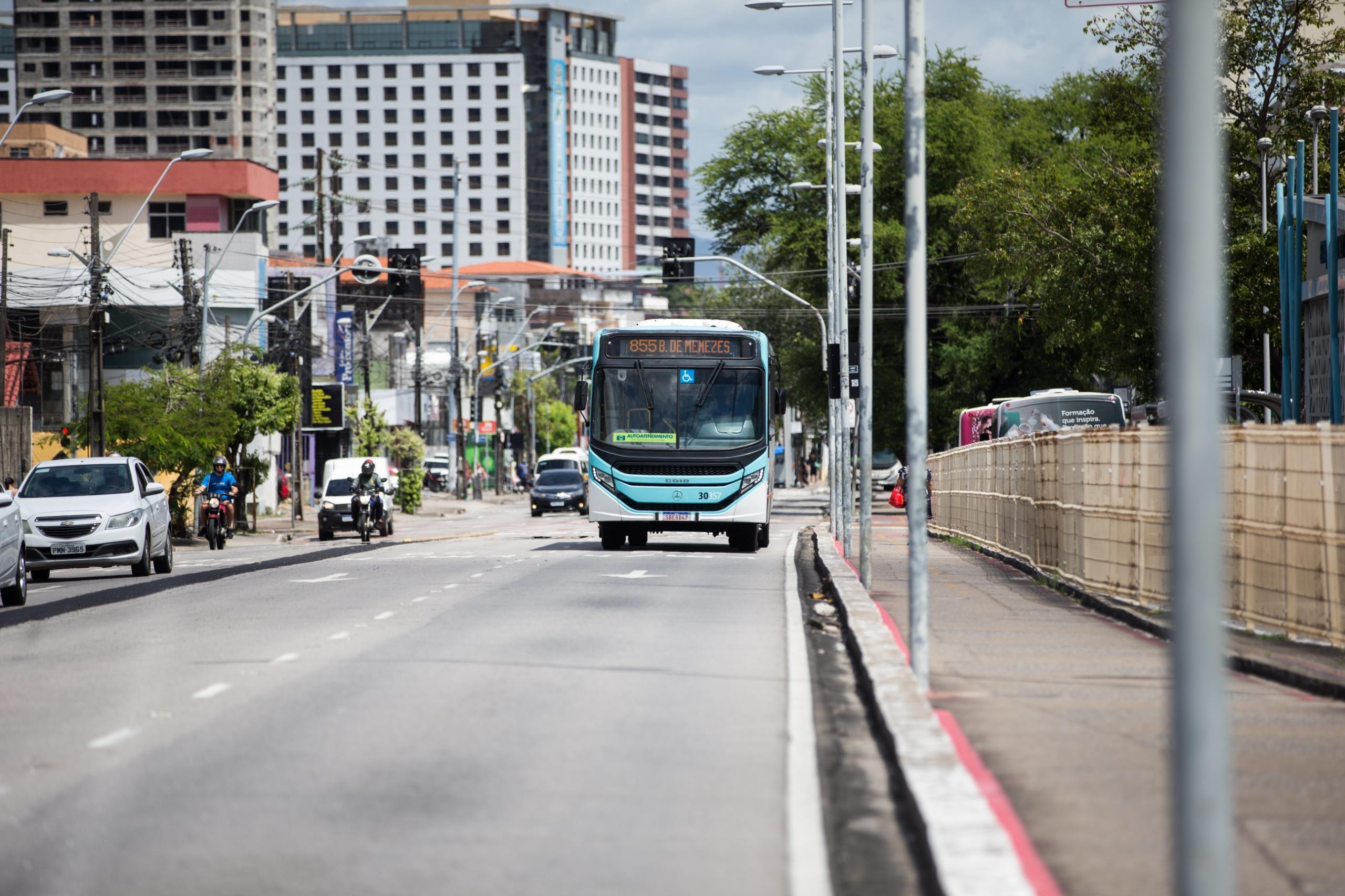 Ônibus urbano azul e branco em movimento numa avenida movimentada, com carros, edifícios e árvores ao fundo sob sol forte.