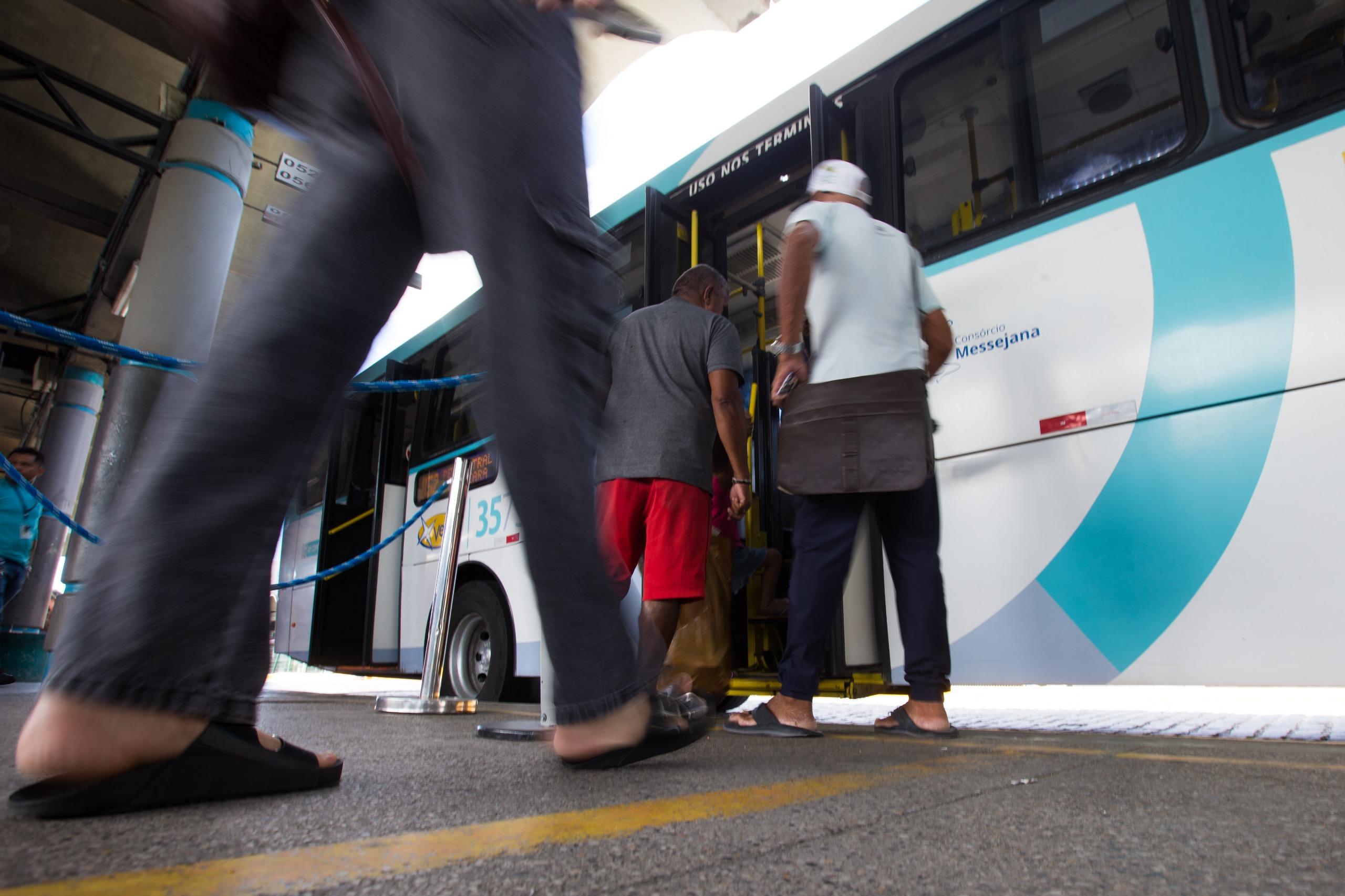 Pessoas embarcando em um ônibus branco e azul em um terminal de transporte público, com passageiros em movimento visíveis da cintura para baixo.