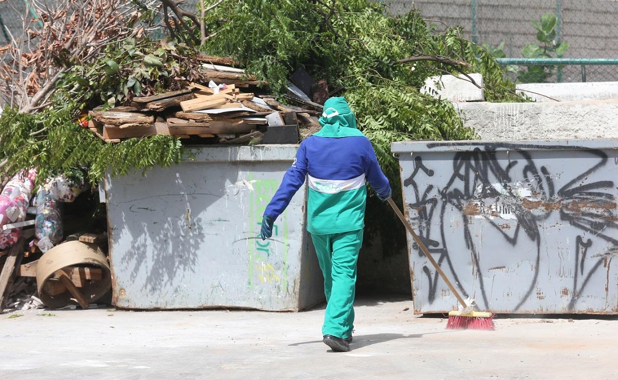 Um gari de Fortaleza, vestido com uniforme azul e verde, varrendo o chão em frente a contêineres de lixo cheios de galhos e detritos de madeira, em um dia ensolarado num Ecoponto.