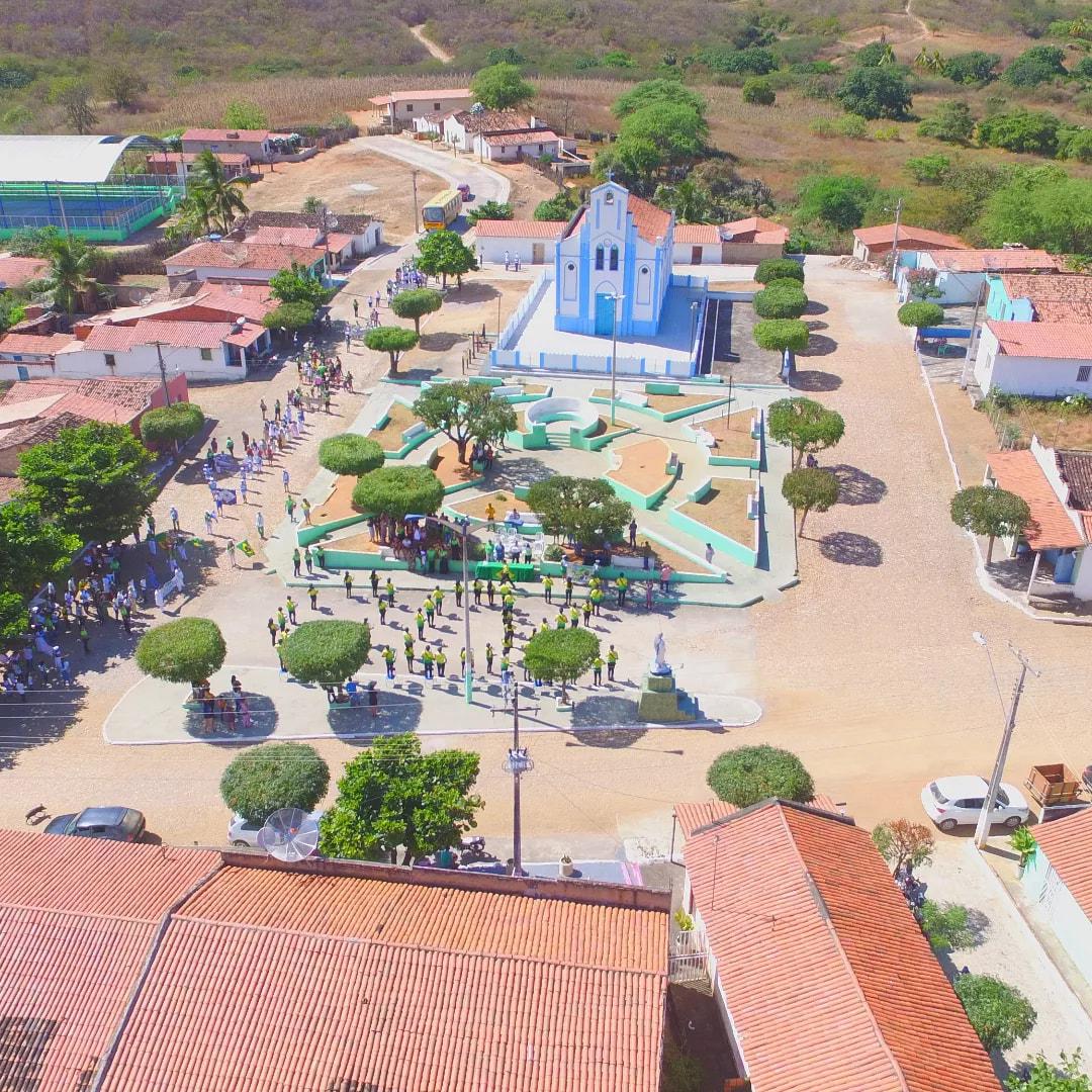 Vista aérea de Itatira, no Ceará, com uma igreja azul e branca no centro de uma praça movimentada, cercada por casas de telhado vermelho e colinas verdes ao fundo.