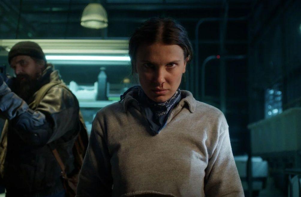 Eleven (Millie Bobby Brown) com expressão séria e determinada, vestindo um moletom cinza e bandana azul em um ambiente escuro.