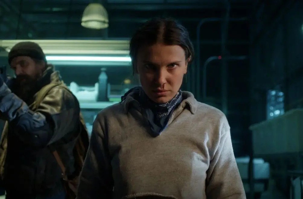 Eleven (Millie Bobby Brown) com expressão séria e determinada, vestindo um moletom cinza e bandana azul em um ambiente escuro.