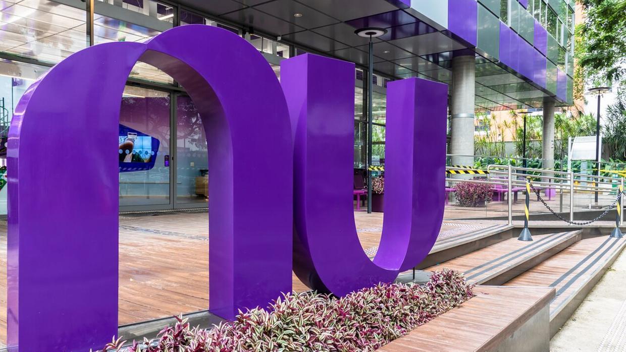 Escultura roxa com as letras “nu” em frente a um prédio moderno com fachada de vidro e área externa com plantas.