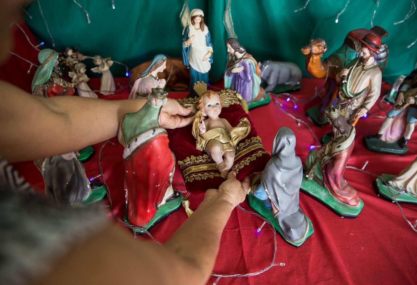 Mãos posicionando a figura do Menino Jesus em um presépio de Natal, com outros personagens como Maria e os Reis Magos ao redor, sobre um tecido vermelho com luzes.