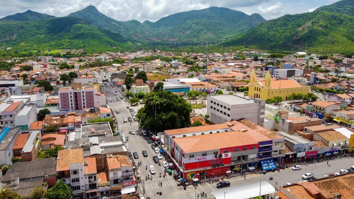 Vista aérea de uma cidade de médio porte, com ruas movimentadas, prédios baixos e telhados alaranjados. Em destaque, uma igreja amarela com duas torres em uma praça central. Ao fundo, há morros e montanhas cobertos por vegetação verde sob um céu parcialmente nublado.