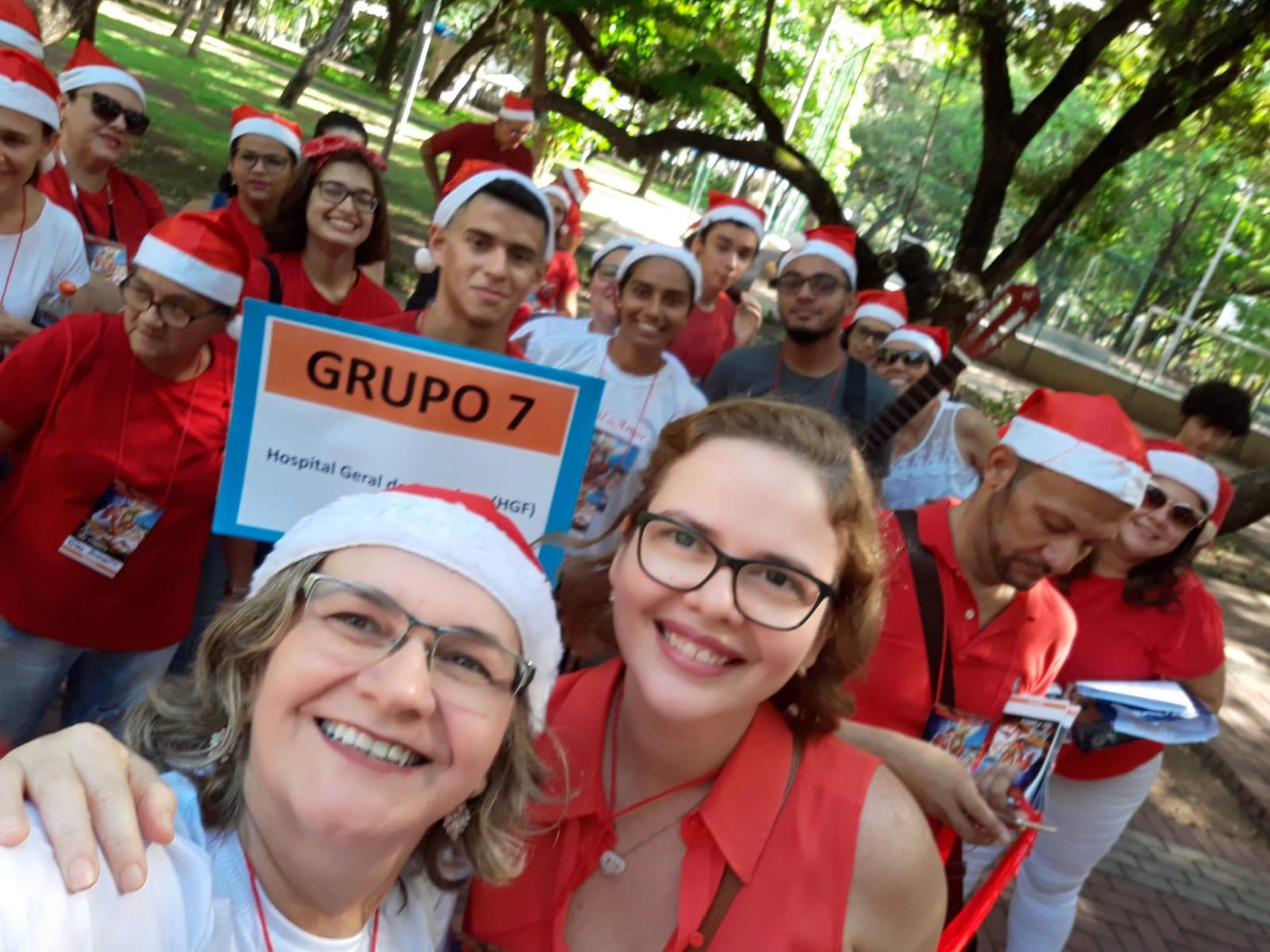 Na imagem, uma fotografia em plano médio, estilo selfie, tirada em um corredor de hospital. Em primeiro plano, uma mulher loira de óculos sorri para a câmera, vestindo uma camiseta branca e um gorro de Papai Noel. Ao fundo, o corredor está repleto de voluntários também usando gorros de Natal e, em sua maioria, camisetas vermelhas. Alguns usam máscaras de proteção facial. À esquerda, uma mulher de vermelho segura sacolas com o que parecem ser presentes ou suprimentos. Mais ao fundo, o grupo se organiza em fila; um homem segura um violão, sugerindo uma apresentação musical ou visita de caridade. O ambiente possui iluminação hospitalar branca, paredes claras e, à direita, é possível ver uma maca metálica estacionada. O clima é de solidariedade e celebração natalina em um ambiente de saúde.