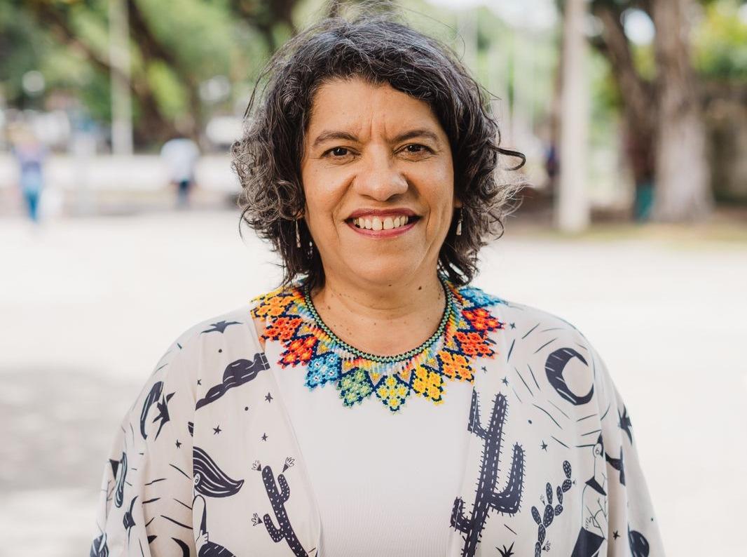 Mulher sorridente, de cabelos curtos e grisalhos, posa ao ar livre em um parque arborizado. Ela usa blusa clara com estampas em preto e um colar colorido de miçangas, olhando diretamente para a câmera.