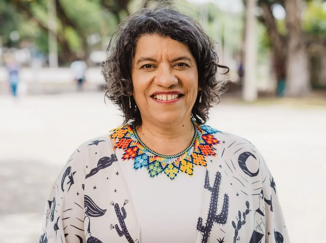 Mulher sorridente, de cabelos curtos e grisalhos, posa ao ar livre em um parque arborizado. Ela usa blusa clara com estampas em preto e um colar colorido de miçangas, olhando diretamente para a câmera.