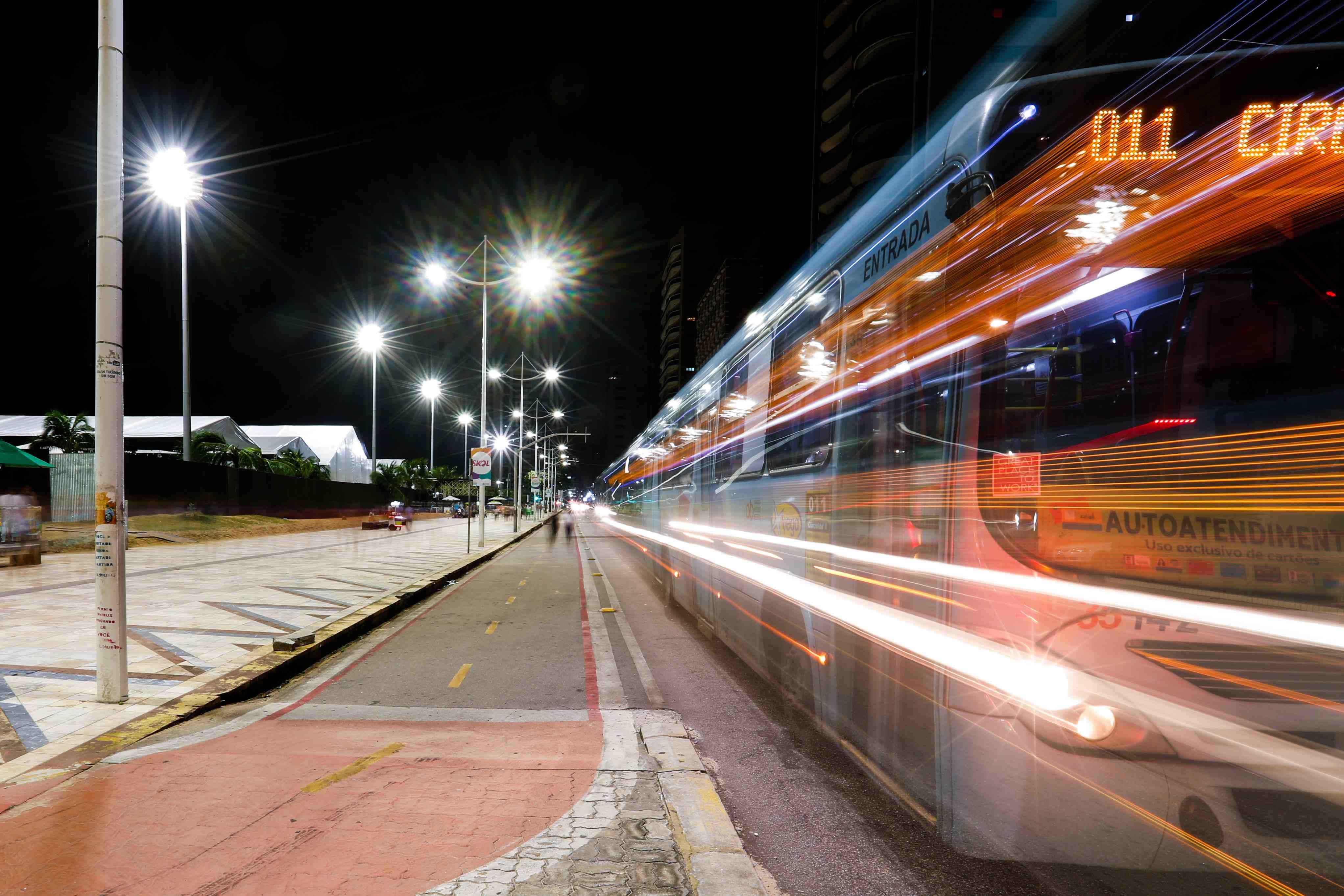 Fotografia noturna com técnica de longa exposição de um ônibus em movimento em uma avenida, criando rastros de luz coloridos ao lado de um calçadão iluminado por postes.