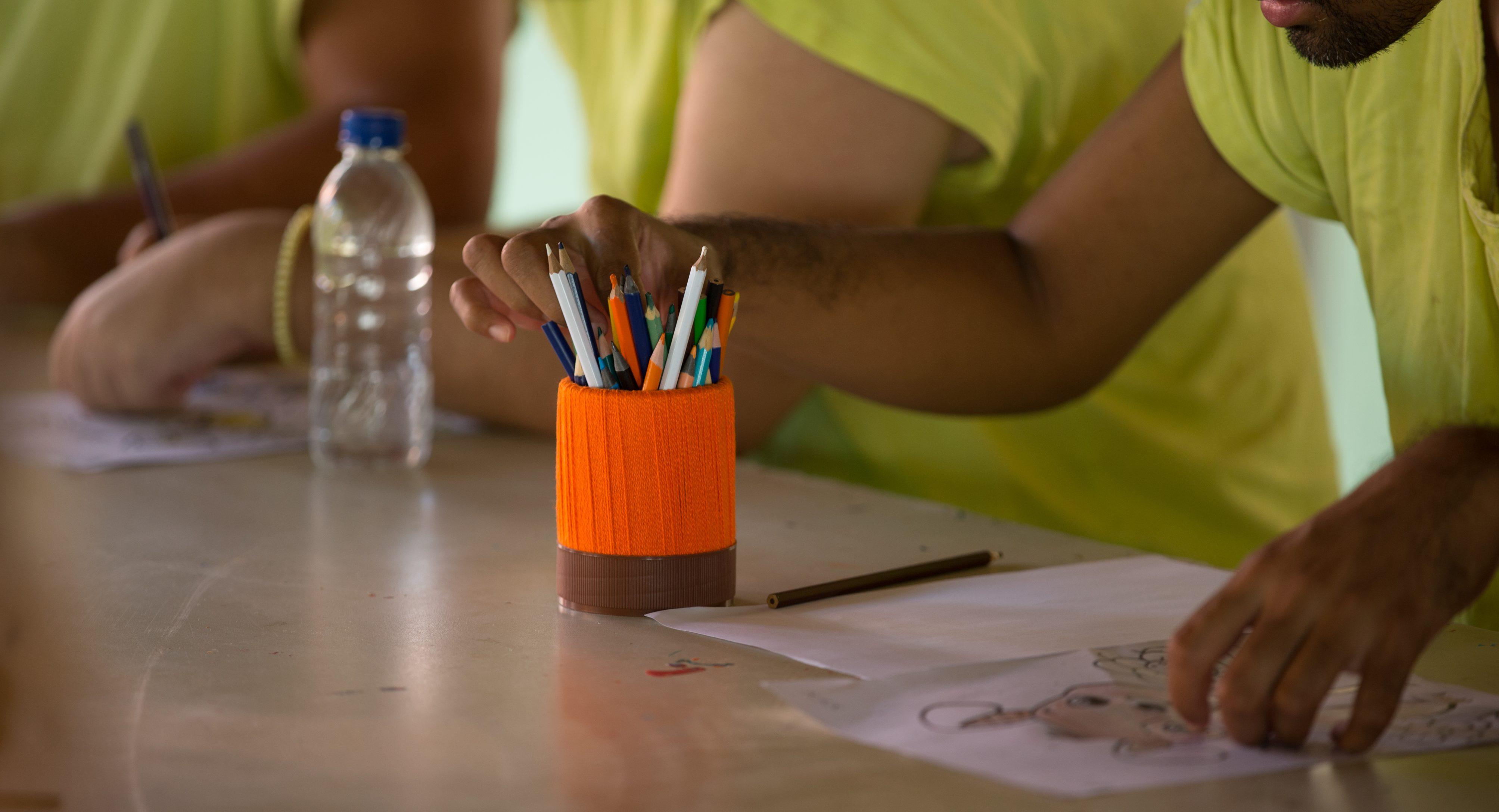 Close-up de mãos de pessoas vestindo camisetas amarelas sentadas ao redor de uma mesa de atividades. Uma mão alcança um lápis de cor em um suporte laranja vibrante, enquanto outras mãos organizam papéis com desenhos para colorir.