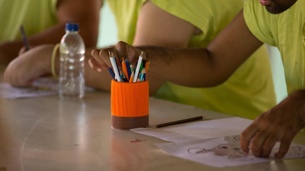 Close-up de mãos de pessoas vestindo camisetas amarelas sentadas ao redor de uma mesa de atividades. Uma mão alcança um lápis de cor em um suporte laranja vibrante, enquanto outras mãos organizam papéis com desenhos para colorir.