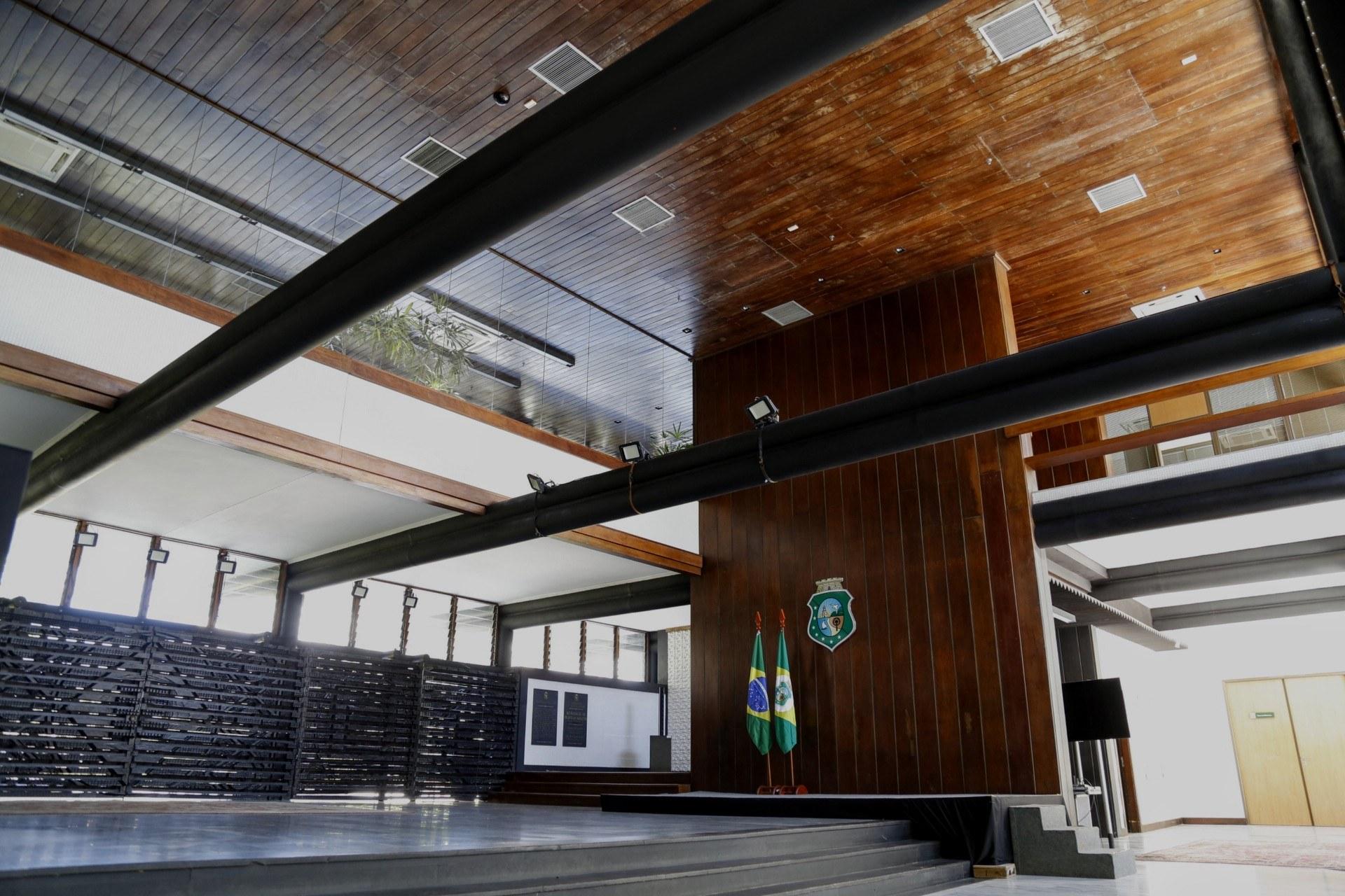 Interior de auditório no Palácio da Abolição, com palco elevado, escudo do Ceará ao centro, bandeiras do Brasil e do Estado, teto e paredes de madeira, vigas metálicas aparentes e luz natural entrando por grandes aberturas laterais.