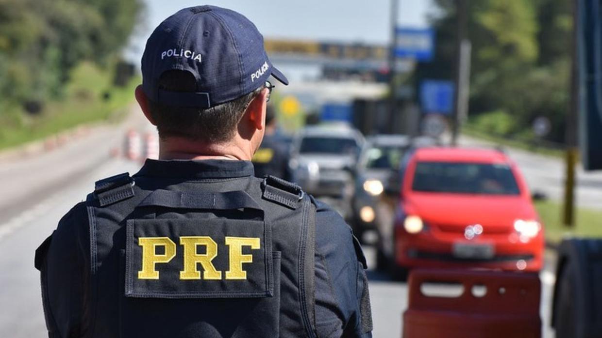 Agente policial com nome PRF escrito nas costas parado e de costas olhando veículos em via.