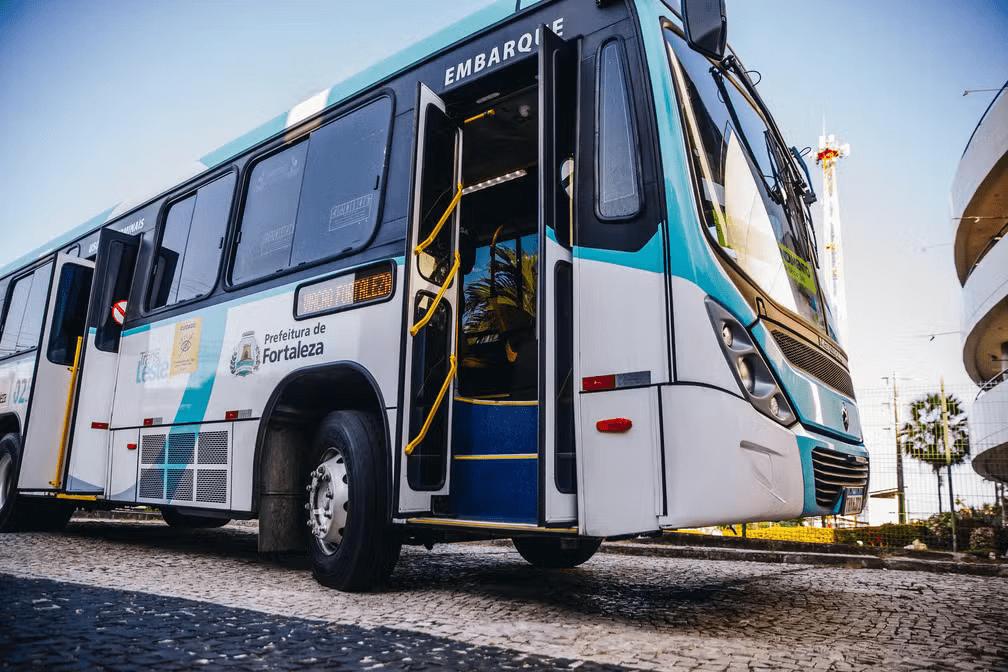 Ônibus urbano branco e azul da Prefeitura de Fortaleza, visto em ângulo baixo, com a porta dianteira aberta e a palavra “Embarque” acima da entrada, parado em uma rua de paralelepípedos sob céu azul.