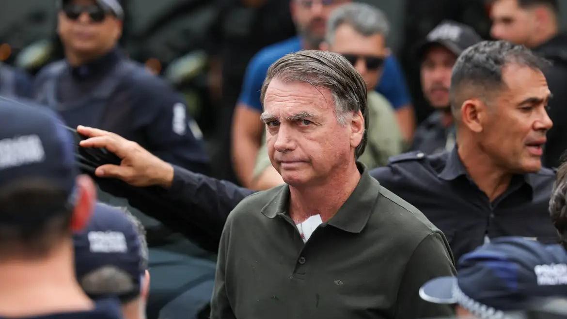 Jair Bolsonaro em meio a seguranças, olha para a câmera enquanto ele entra em um carro. O político veste uma camisa polo. Foto usada em matéria sobre cirurgia do ex-presidente.