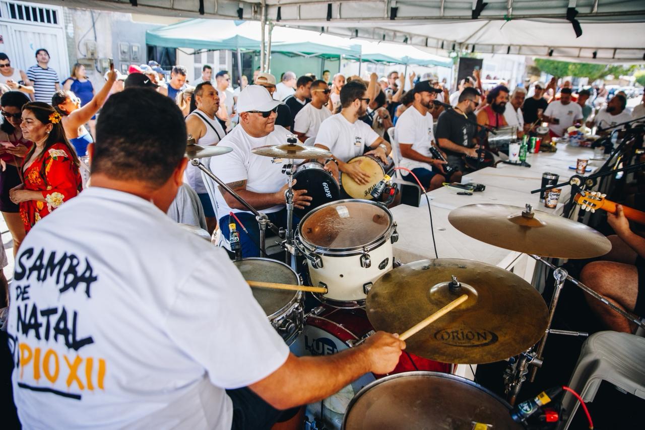 Roda de samba acontece sob uma grande tenda, com músicos sentados em volta de mesas tocando instrumentos de percussão. Em primeiro plano, um baterista toca a bateria; ao fundo, outros integrantes tocam tantan, pandeiro e cavaquinho, enquanto o público acompanha de pé, alguns dançando e batendo palmas, em clima festivo e coletivo.