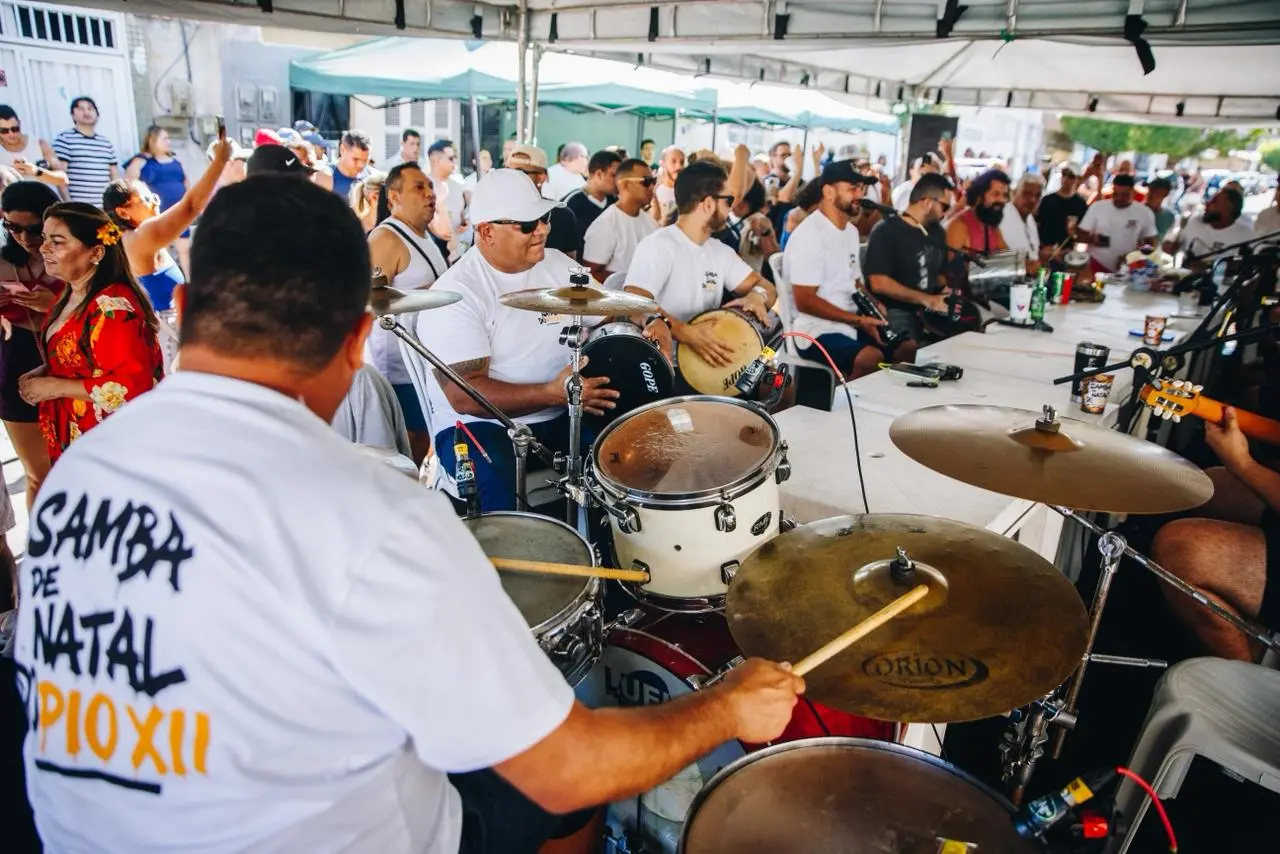 Roda de samba acontece sob uma grande tenda, com músicos sentados em volta de mesas tocando instrumentos de percussão. Em primeiro plano, um baterista toca a bateria; ao fundo, outros integrantes tocam tantan, pandeiro e cavaquinho, enquanto o público acompanha de pé, alguns dançando e batendo palmas, em clima festivo e coletivo.