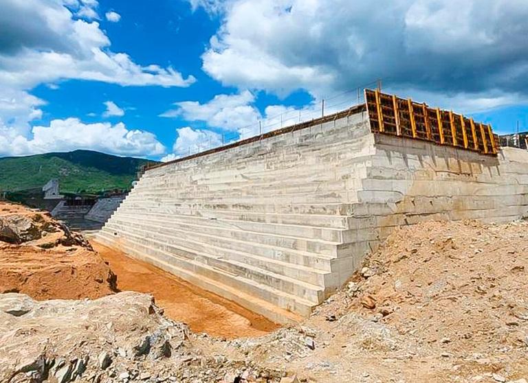 Grande estrutura de concreto em construção, com degraus de contenção e forma de cofragem no topo, sob um céu azul com montanhas verdes ao fundo, de uma barragem no Ceará.