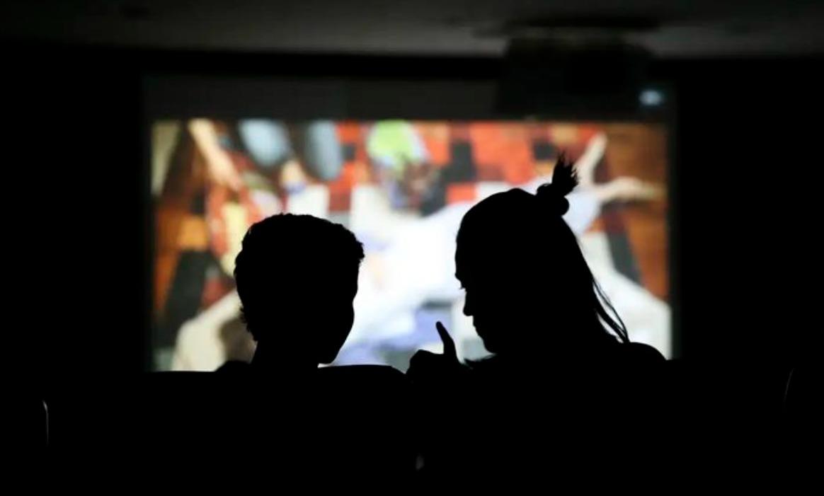 Silhueta em close-up de duas pessoas sentadas em uma sala escura, possivelmente um cinema ou auditório. À esquerda, vê-se o perfil de uma criança e, à direita, o perfil de um adulto com o cabelo preso em um coque, que parece gesticular com a mão enquanto conversa com a criança. Ao fundo, uma tela de projeção brilhante exibe uma imagem colorida e levemente desfocada, criando um contraste acentuado com as figuras em primeiro plano.