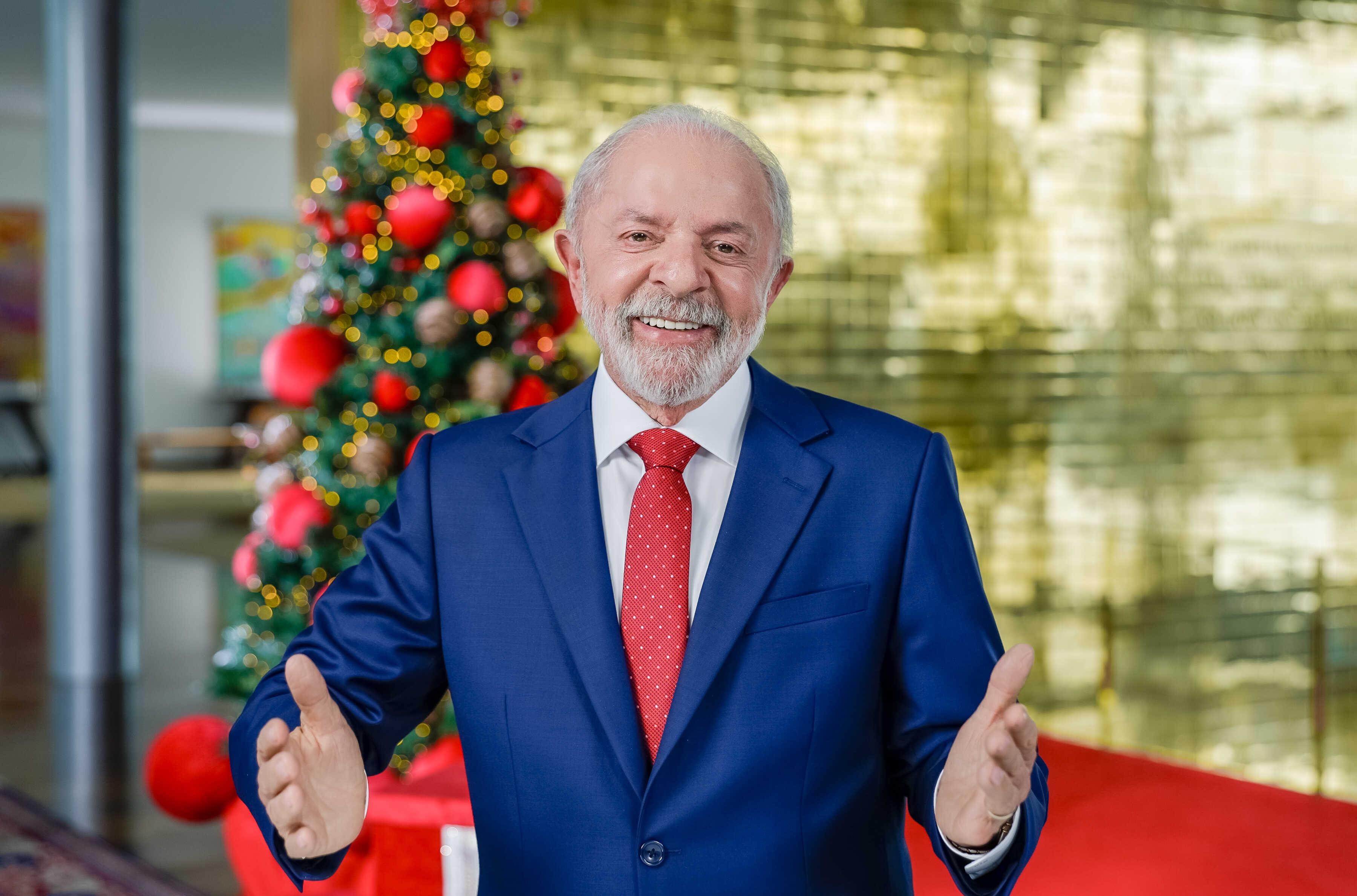 Fotografia do presidente do Brasil, Luiz Inácio Lula da Silva, sorrindo de braços abertos em um ambiente festivo. Ele veste um terno azul-marinho, camisa branca e gravata vermelha com pequenos detalhes brancos. Ao fundo, à esquerda, há uma árvore de Natal decorada com bolas vermelhas e douradas.