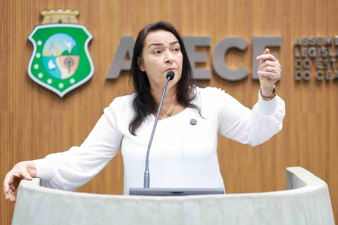 A imagem mostra uma mulher de meia-idade com cabelos escuros, vestindo uma camisa social branca de manga longa, falando em um púlpito. Ela está de pé, com o microfone perto do rosto, e gesticula com a mão direita levantada. Ao fundo, há um painel de madeira com o emblema do estado do Ceará à esquerda e o texto parcial 
