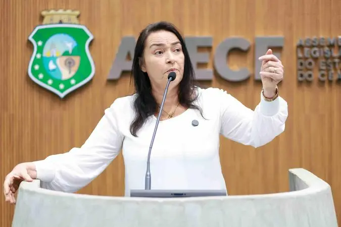 A imagem mostra uma mulher de meia-idade com cabelos escuros, vestindo uma camisa social branca de manga longa, falando em um púlpito. Ela está de pé, com o microfone perto do rosto, e gesticula com a mão direita levantada. Ao fundo, há um painel de madeira com o emblema do estado do Ceará à esquerda e o texto parcial 