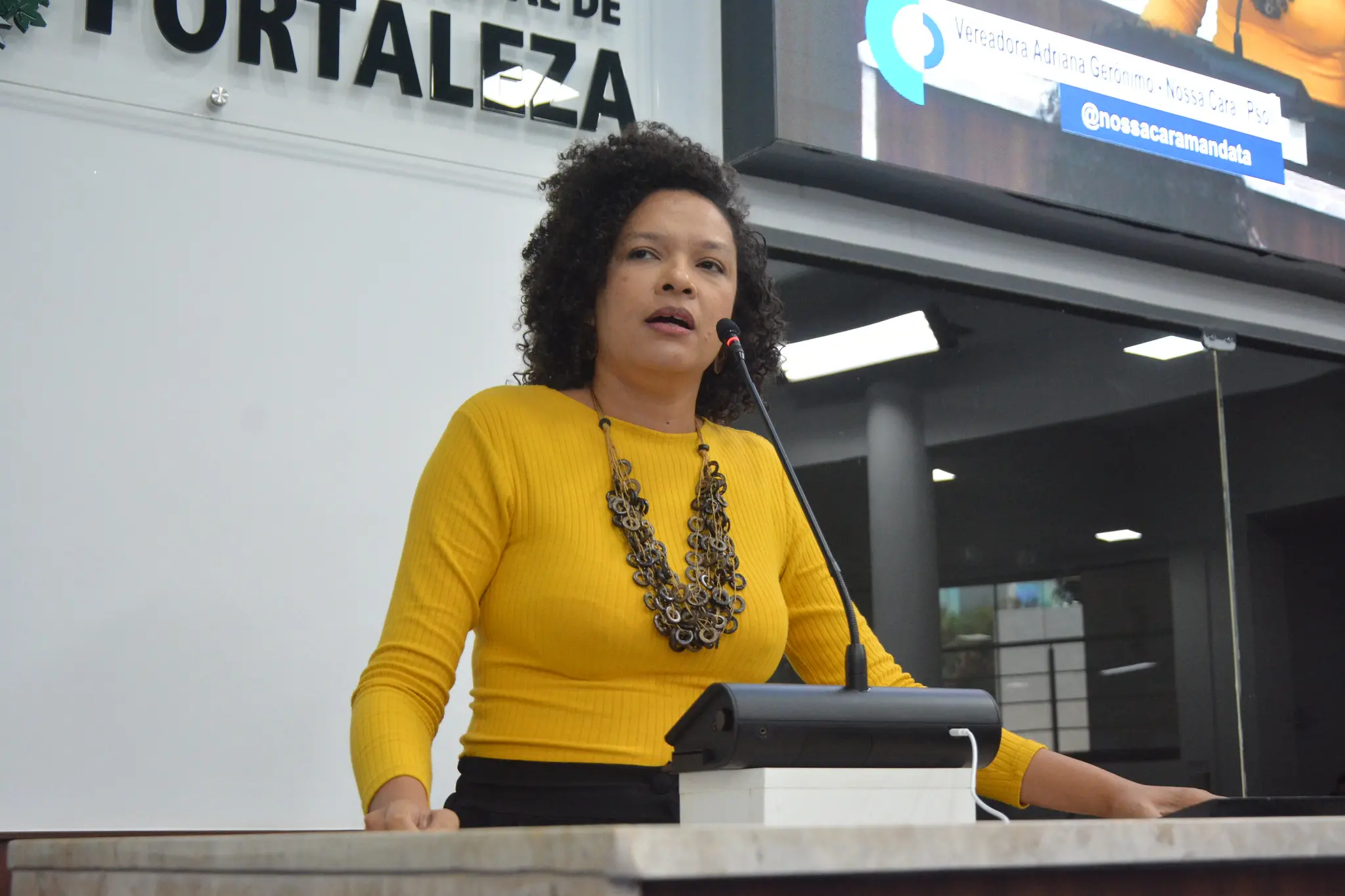 Foto que contém Adriana Gerônimo, vereadora de Fortaleza pelo Psol. Ela é uma mulher negra de cabelo cacheado longo voltado para trás, usa um colar e veste uma camisa de mangas longas amarelas.