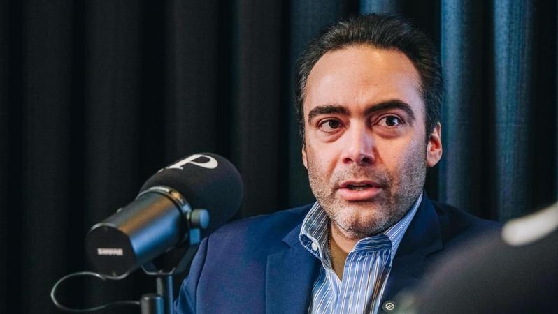 A imagem mostra um homem de terno azul e camisa social listrada em tons claros, falando diante de um microfone preto com a letra “P” em destaque na espuma protetora. Trata-se de Ozires Pontes. Ele tem pele clara, cabelos curtos e escuros, penteados para trás, além de barba cerrada. Ao fundo, há uma cortina escura desfocada, sugerindo que a cena ocorre em um estúdio de gravação ou entrevista.