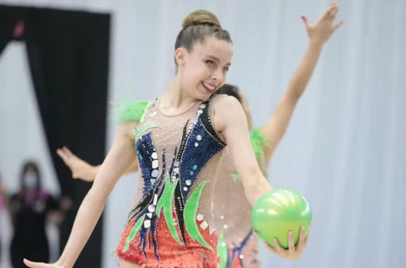 Isabelle Marciniak com bola verde claro nas mãos, coque alto na cabela e maiô com as cores azul e laranja.
